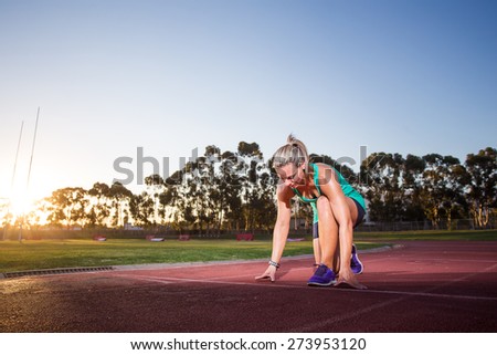 Close up wide angle view of a female sprinter  athlete getting ready to start a race on a tartan racetrack with dramatic lighting late in the afternoon, just before dusk.