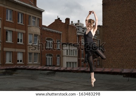 Female ballet dancer in black dress performs pointe rotation with her arms closed above the head on a rooftop in a city