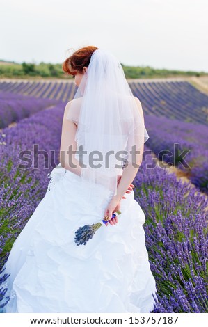 A young European woman in a wedding dress stands back, a hand bouquet of lavender, lavender field on background