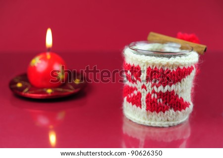 A mug of tea in the knitted cover with a winter ornament, cinnamon stick, lit a candle on red background