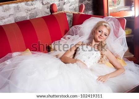 beautiful  bride wearing white wedding dress and veil lying on red sofa in interior of hall
