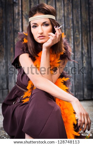 beautiful actress in brown and orange boa smoking against old wooden gate