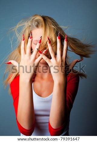 blonde girl with hair fluttering on wind hiding her face by beautiful hands with long fingers and fashionable design of nails on dark blue background