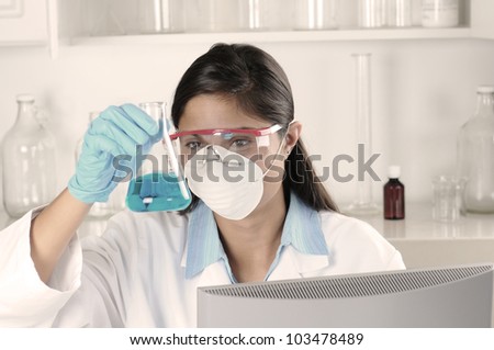 Young female lab tech seated behind computer monitor holding flask and inspecting contents. Horizontal format.