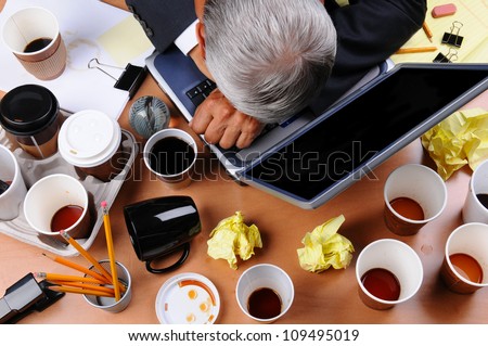Closeup view of a very cluttered businessman\'s desk. Overhead view of the mature man\'s head on laptop keyboard and scattered coffee cups and office supplies. Horizontal format.