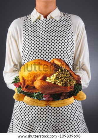 Closeup of a homemaker in an apron and oven mitts holding a platter with a roasted Thanksgiving turkey. Horizontal format over a light to dark background. Woman is unrecognizable.