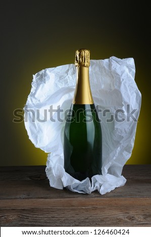 Still life of champagne bottle wrapped in tissue paper on wood surface and light to dark background.