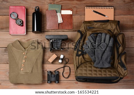 Overhead view of hiking gear laid out for a backpacking trip on a rustic wood floor. Items include, Backpack, gloves, sweater, camera, film, binoculars, passport, wallet, canteen, compass, money,