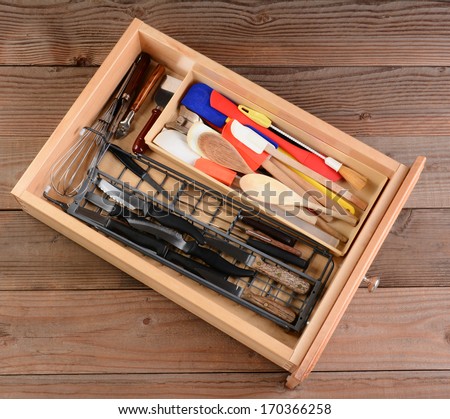 High angle view of a kitchen drawer on a rustic wooden table. The drawer is full of wooden spoons, knives, whisks spatulas, and other household kitchen items.