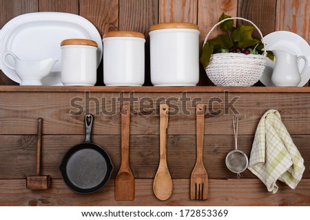 Closeup of a rustic kitchen wall. One shelf with canisters, plates and a basket. Hanging on the wall below are wooden utensils, frying pan and towel.