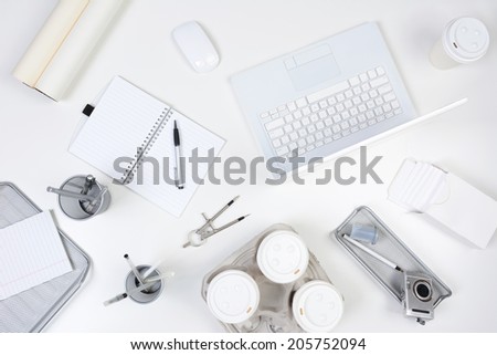 High angle shot of a white desk with primarily white and silver office objects. Items include, laptop computer, pad, pens, coffee cups, compass, camera and film.