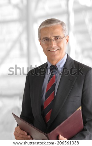 A mature businessman with a leather folder in a modern office building. The background is out of focus and high key.