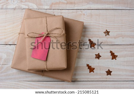 A high angle view of plain brown paper wrapped presents with red tags on a white rustic table. Small rusty metal ornaments in star and tree shapes lay by their side. Horizontal format.