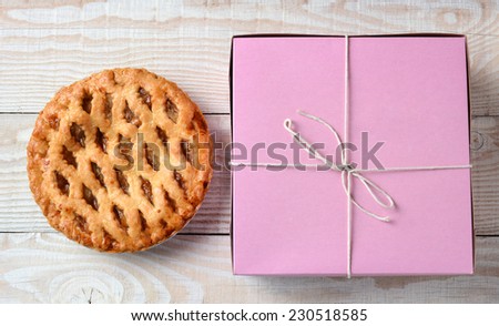 High angle shot of an apple pie and a pink bakery box. Horizontal format on a rustic white wood kitchen table.