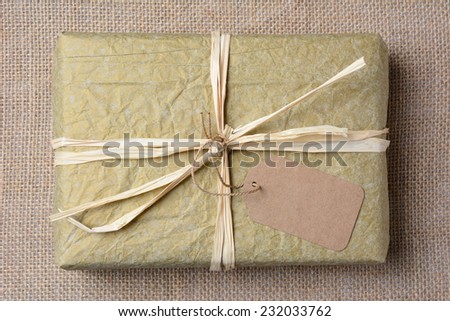 Closeup of a gold tissue paper wrapped present on a burlap surface. The gift is tied with raffia and a blank git tag. High angle shot in horizontal format.