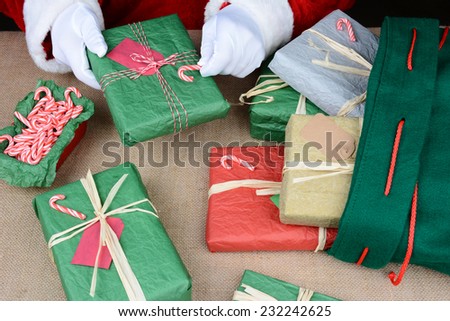 Closeup of Santa Claus as he puts the finishing touches on presents before stuffing them into his bag. Horizontal format showing only the jolly old elfs hands as he places candy canes on a gift.