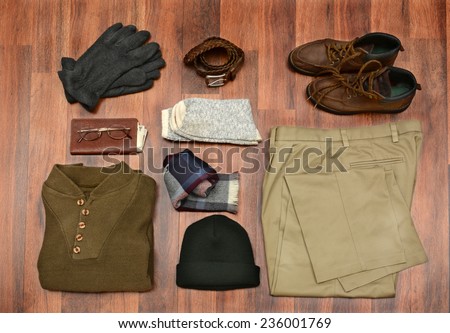 High angle shot of men\'s winter clothes laid out on a dark wood floor. Items include, Sweater, Scarf, Gloves, wool Socks, Pants, Boots, belt, Knit Cap, Wallet, and Glasses. Horizontal Format.