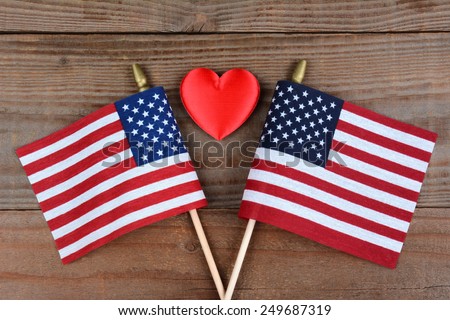 High angle shot of two crossed American flags on a rustic wood surface with a red heart inbetween. Horizontal format.