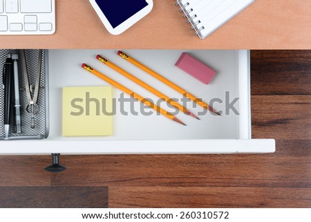 High angle shot of an open desk drawer showing the items inside. The top of the desk has a computer keyboard Cell Phone and note pad. The neat drawer has pencils paper and organizer.