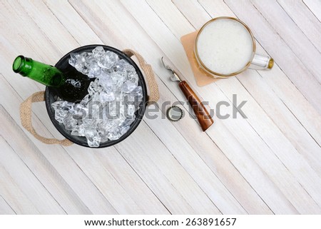 Overhead shot of an ice bucket with an opened beer bottle, a mug of beer and opener on a rustic white wood table. Horizontal format with copy space.