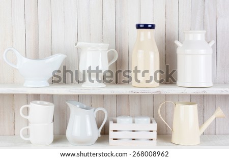 Closeup of two white wood rustic kitchen shelves with assorted household items. Various objects, include, salt and pepper shakers, cups, pitchers bottles and crockery.