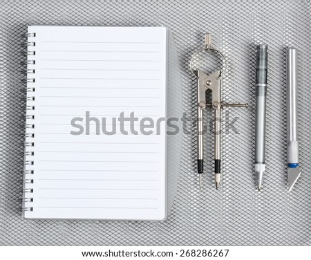 High angle shot of an open notebook with pen, compass and razor on a silver mesh background.