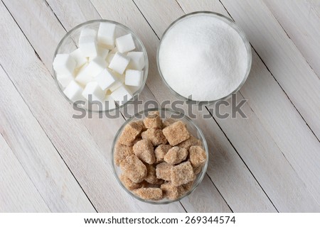 Three glass bowls filled with granulated sugar, white sugar cubes and natural sugar chunks. High angle view on a rustic wood kitchen table.