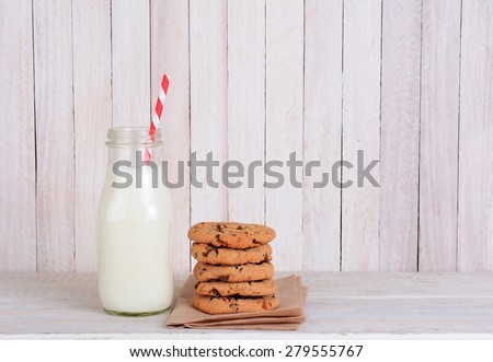 An after school snack of cookies and milk on a rustic wood set. A small bottle of milk with a drinking straw and a stack of chocolate chip cookies. Horizontal format with copy space.