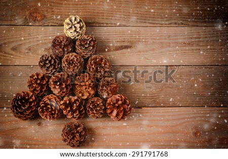A group of pine cones arranged in a Christmas Tree shape on a rustic wood floor with snow flakes effect. Horizontal format with an instagram retro look and copy space.