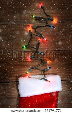 High angle shot of a Christmas stocking on a rustic wood surface with holiday lights sticking out the top. Vertical format with snow flakes effect.