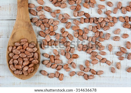 Overhead view of a wood spoonful of pinto beans surrounded by more loose beans. Horizontal format on a rustic whitewashed wood table.