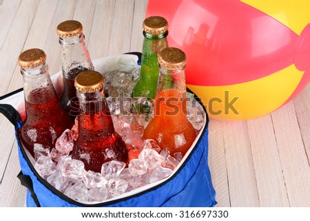 Closeup of a cooler with soda bottles and ice on a rustic wood table with a beachball in the background.