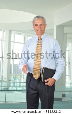 Middle Aged Businessman with notebook and hand extended to shake in modern office setting