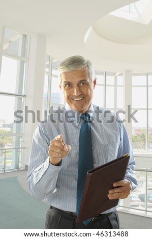 Smiling Middle Aged Businessman Holding Folder and Pointing at the camera in modern office setting
