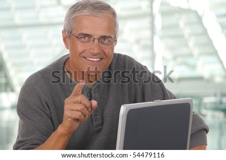 Middle aged man sitting at laptop computer and pointing at camera. Man is smiling and casually dressed. Horizontal format with blurred office background.