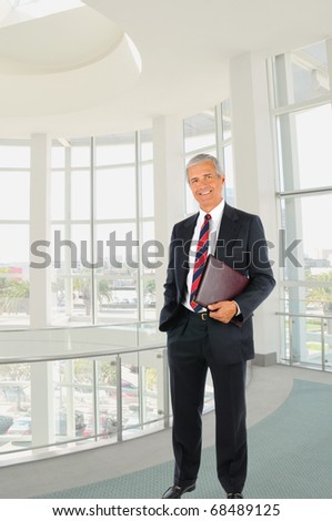 Smiling middle aged businessman in a suit and tie standing in a modern office building with one hand in his pocket and the other holding a leather folder.