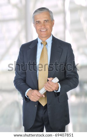 Middle aged businessman in a suit and tie standing in a modern office building holding a rolled up newspaper. Vertical Format