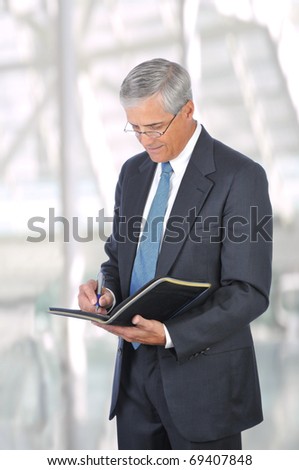 Middle aged businessman in a suit and tie standing in a modern office building writing in a notebook. Vertical Format