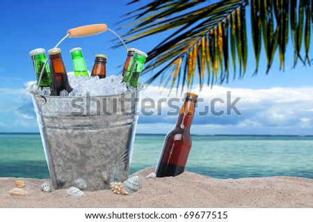 Assorted beer bottles in a bucket of ice in the sand on a tropical beach. One beer bottle without a cap is by itself stuck in the sand next to the pail.
