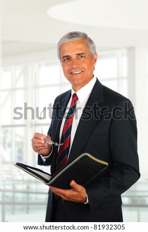 Smiling middle aged businessman holding a binder and his eye glasses while standing in an office lobby.