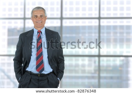 Smiling middle aged business man isolated on white with his hands in pants pockets. Vertical Format standing in front of large window of modern office building.