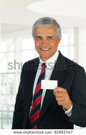 Smiling middle aged businessman in modern office setting holding out blank business card. Vertical Format.