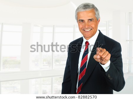 Middle aged businessman in modern office setting pointing at camera. Horizontal Format.