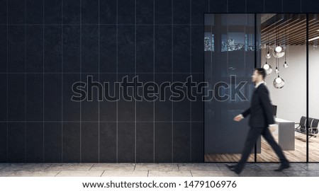 Businessman walking past dark wall business center with modern light conference room with wooden floor and black chairs.