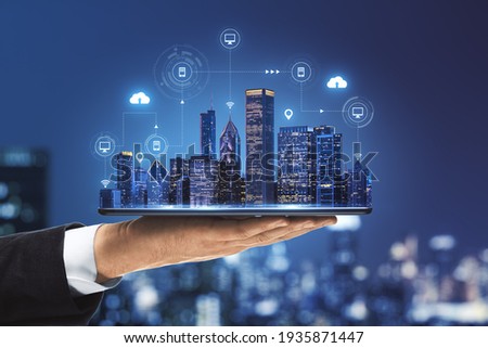 Smart city concept with real skyscrapers layout with glowing digital cloud technology icons on digital tablet screen that carrying businessman hand on blurry megapolis city background
