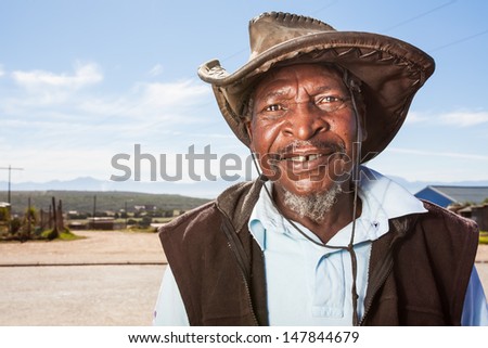 image of an old african man wearing a leather farming hat standing outside smiling with watery eyes