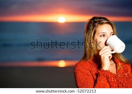 Pretty woman wearing a red wool top drinking a cup of coffee on the beach at sunrise.