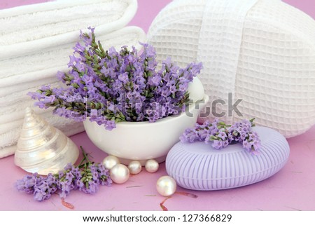 Lavender herb flower sprigs, soap, white towels and body rub with pearls and shells over mottled lilac background.