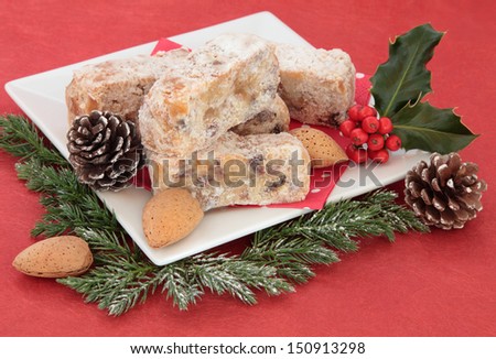 Stollen christmas cake slices with holly, almond nuts, pine cones and winter greenery decorations over red background.