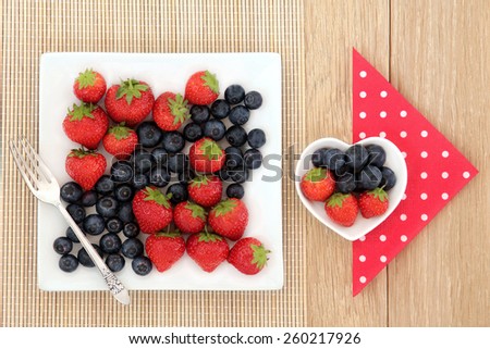 Strawberry and blueberry fruit in square and heart shaped porcelain dishes with polka dot napkin and old silver fork over oak and bamboo.